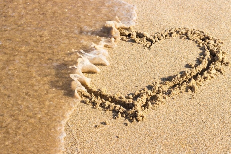 Hart getekend in het zand dat door een golf wordt weggevaagd – symbool voor een onverwachte scheiding en het verlies van een relatie.