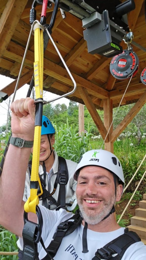 Rinaldo glimlacht bij de start van de zipline op Madeira, met helm en veiligheidsharnas onder een houten overkapping.