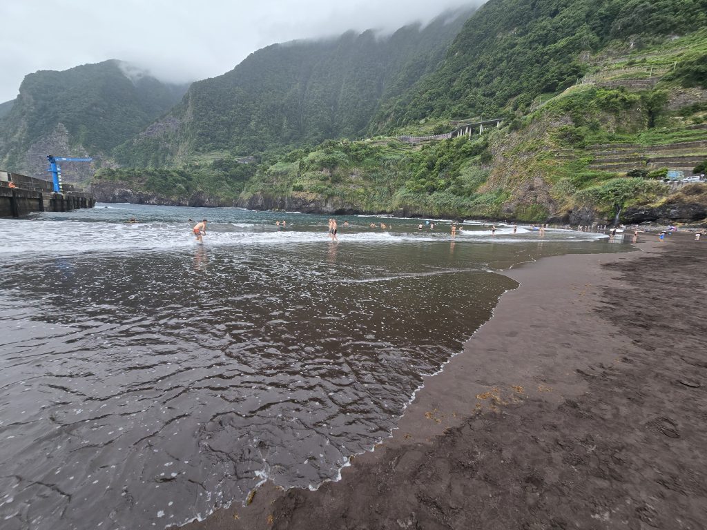 Badgasten op het donkere zandstrand van Seixal met terrassen en groene berghellingen.