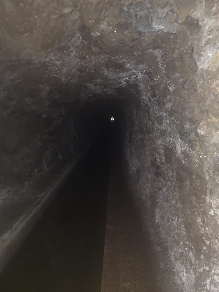 Donkere levadatunnel met watergoot in Madeira, enkel verlicht door een klein lichtpunt aan het einde op de PR16.